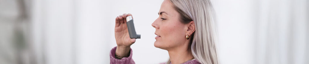 Sie sehen eine Frau mit einem Inhalator in der Hand. Sie leidet an Bronchialasthma. Leitsymptom von Asthma bronchiale (Bronchialasthma) ist die anfallsartig auftretende Atemnot. Schleimhautschwellung, Sekret und Verkrampfung verengen die Atemwege. Der kleiner gewordene Durchmesser der Luftwege erschwert die Einatmung, vor allem aber die Ausatmung. Der Anfall beginnt mit plötzlich einsetzender Luftnot und einem zunehmenden Engegefühl in der Brust, das von Husten begleitet werden kann. Die Einatmung geht in der Regel leichter als die Ausatmung.